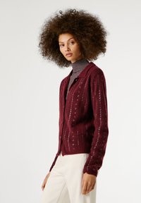 Κάρντιγκ μπορντό πλεκτό με κατακόρυφες ραφές, κυκλικά κουμπιά και ανοιχτή ύφανση. Στυλιζαρισμένο πάνω από ένα ριγέ ζιβάγκο.