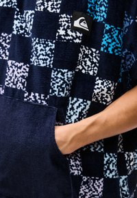 Χέρι στην τσέπη ενός ναυτικού μπλε και σχεδιασμένου κοντομάνικου φούτερ με κουκούλα, με εικονοστοιχειωτό σχέδιο σκακιέρας σε ανοιχτό μπλε και λευκό και με μπαλωμένο λογότυπο Quiksilver.