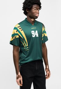 Πράσινη αθλητική φανέλα με κίτρινες και τιρκουάζ ρίγες, αριθμό 94 και λογότυπο adidas. Κοντά μανίκια και άνετη ε φαρμογή. Μαύρο τζιν παντελόνι.