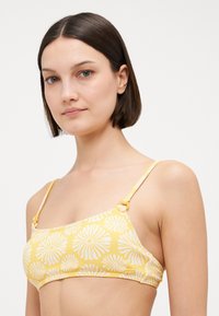 Žltý bikini top s kvetinovým vzorom, tenkými ramienkami a textúrovanou látkou. Obsahuje biele potlače pripomínajúce margarétky a nastaviteľné hardvérové ramienka.