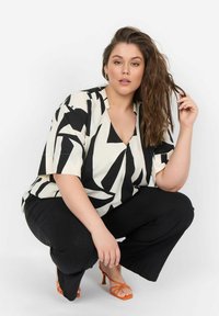 Zwart-wit abstract patroon korte mouwen top, V-hals, gecombineerd met zwarte broek en oranje sleehakken, wat de gedurfde contrasten benadrukt.