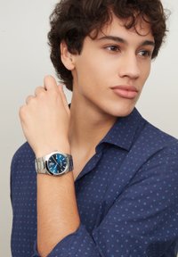 Orologio in acciaio inox argentato con quadrante blu, caratterizzato da un design semplice e un bracciale liscio, indossato su un polso con una camicia blu a fantasia.