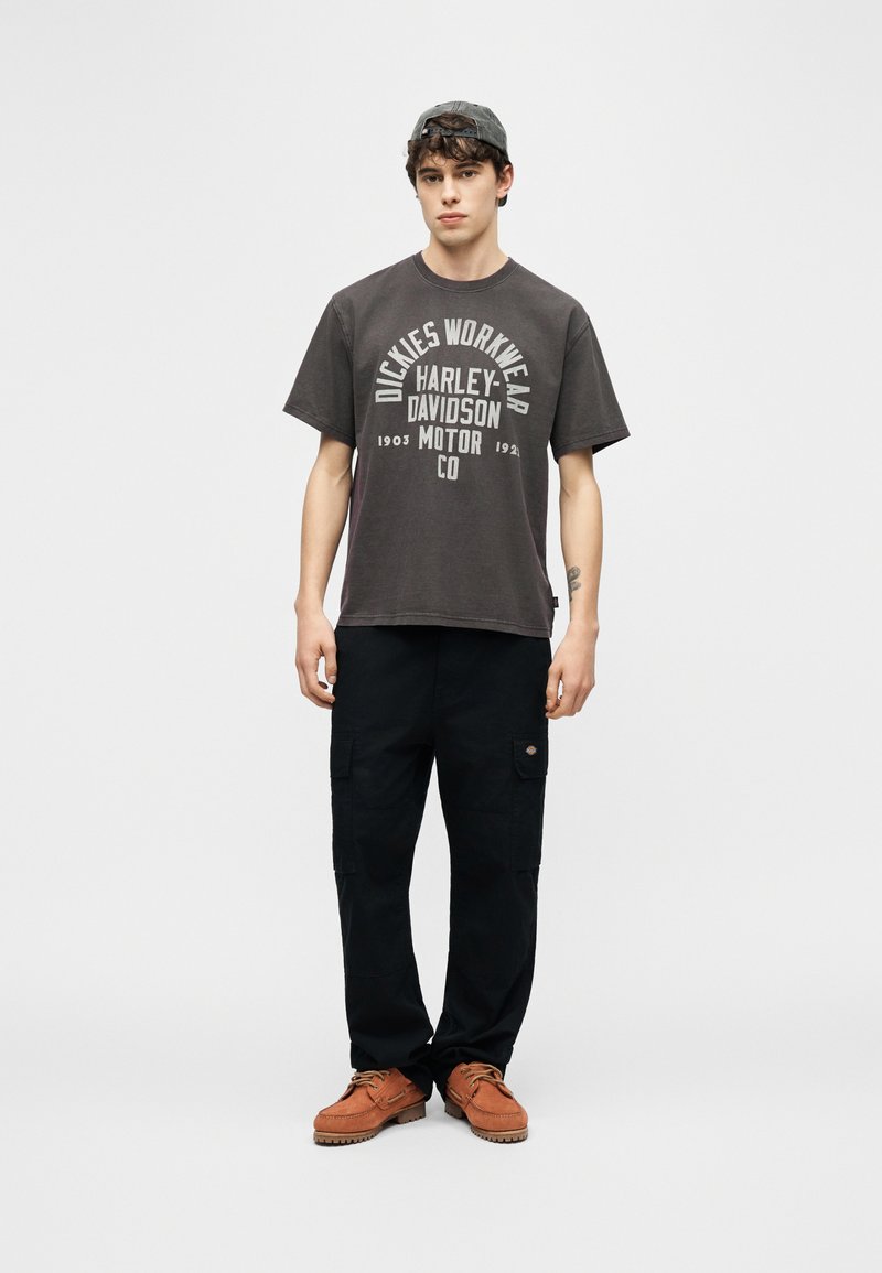 Giovane uomo che indossa una t-shirt nera Harley-Davidson Dickies Workwear, pantaloni cargo neri, scarpe marroni e un cappellino grigio indossato al contrario, in piedi su uno sfondo bianco.