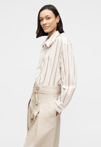 Ριγέ πουκάμισο με κουμπιά σε απαλές παστέλ αποχρώσεις και μακριά μανίκια, συνδυασμένο με μπεζ παντελόνια ψηλόμεσα που διαθέτουν ζώνη δέσιμου στη μέση.