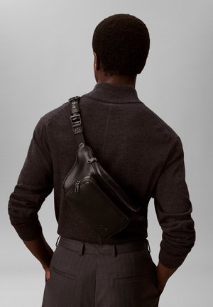 Person iført mørkebrun sweater og grå bukser med striber, med sort læder crossbodytaske båret over ryggen mod en ensfarvet baggrund.