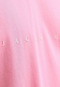 Camiseta de algodón rosa con letras blancas en relieve que deletrean "JACK & JONES". La textura es suave y lisa, con un diseño sencillo.