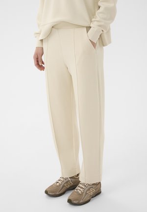 Personne portant un pantalon droit couleur crème avec des poches latérales et des baskets beige New Balance, debout devant un fond clair uni.