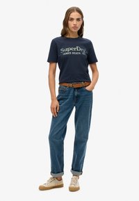 Marineblå bomulds t-shirt med "SuperDry" logo, parret med højtaljede blå denimjeans og tan sneakers. En brun bælte fremhæver outfittet.