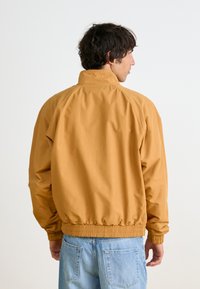 Giacca a vento gialla senape con colletto alto, polsini ed elasticità in vita. Tessuto liscio, con una vestibilità rilassata. Vista posteriore mostrata.