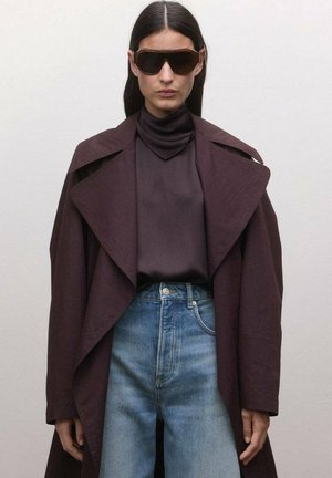 Kobieta ubrana w za duży, ciemnofioletowy płaszcz, top z wysokim kołnierzem, niebieskie dżinsy i duże brązowe okulary przeciwsłoneczne, stojąca na tle jednolitego jasnego tła.