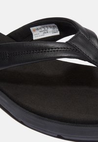Black leather flip-flop with a textured inner sole, featuring soft straps and a rubber outsole for traction. Attached label visible.