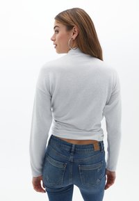 Ljusgrå långärmad topp med en figurnära design, featuring en hög krage och en knut vid midjan, ihop med mörkblå jeans.