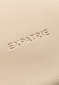 Bežový texturovaný materiál s embosovaným logem "EXPATRIÉ". Povrch má jemnou, zrnitou strukturu typickou pro syntetickou kůži.