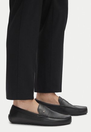 Schwarze Leder-Slipper mit einem durchbrochenen Design, flacher Sohle, runder Zehenform und einem kleinen Logo-Akzent. Kombiniert mit schwarzen maßgeschneiderten Hosen.