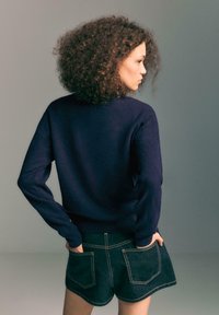 Mornarska pletena majica s dugim rukavima, koja ima okrugli izrez i rebrasti rub, u kombinaciji s tamnim traper šortsevima s žutim šavovima.