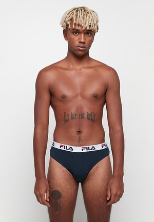 Nuori mies, jolla on vaaleat rastat, seisoo yksiväristä taustaa vasten, päällään tummansinisiä FILA-alushousuja ja näyttäen vatsan ja reiden tatuointeja.