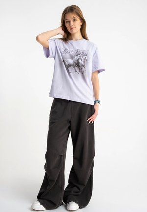 Młoda kobieta stojąca, jedną ręką dotykająca włosów, ubrana w jasnofioletowy t-shirt z grafiką konia, szerokie czarne spodnie i białe trampki.