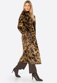 Kabát z umělé kožešiny s leopardím potiskem, dlouhý, se šálovým límcem. Hnědé tóny s černými skvrnami. Nosený přes hnědé slim kalhoty a boty na podpatku.