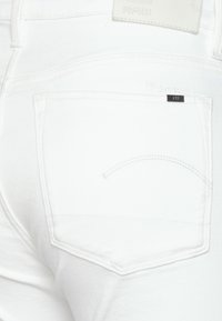 Los vaqueros de mezclilla blanca presentan un bolsillo trasero con un detalle de costura decorativa y una pequeña etiqueta negra que dice "RAW". La tela es suave y ajustada.