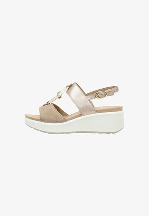 Sandalia de cuña beige y metálica con correa ajustable en el tobillo y cadena decorativa en la parte delantera sobre suela blanca.