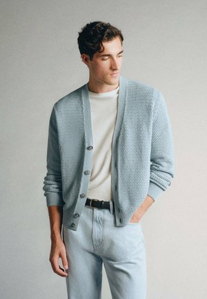 Jeune homme portant un cardigan bleu clair texturé sur une chemise blanche et un jean bleu clair, debout devant un fond uni clair.