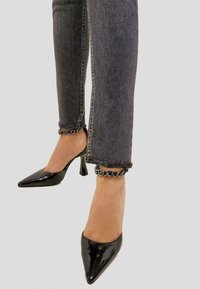 Zapatos de tacón alto de charol negro con puntera afilada, combinados con unos vaqueros de mezclilla gris que presentan un detalle de cadena en el bajo.