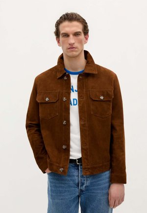 Bruine suède jas met twee borstzakken, knoopsluiting en puntkraag, gedragen over een wit shirt met blauwe accenten. Blauwe spijkerbroek.