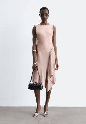 Woman in a sleeveless light pink asymmetric midi dress, white strappy heels, sunglasses, silver jewelry, holding a small black handbag.