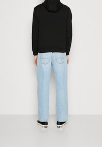 Osoba v svetle modrých rovně střižených džínách, černé mikině s kapucí a černých nízkých teniskách stojí proti jednoduché bílé zdi.