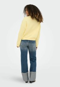 Femme aux cheveux bouclés portant un sweat-shirt jaune et un jean à revers large bicolore, debout de dos sur un fond uni.
