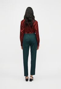 Blouse en satin bordeaux à manches longues, associée à un pantalon vert ajusté. La tenue présente une taille haute et une coupe épurée. Des talons noirs viennent compléter le look.