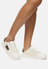 Sneaker bianche con pannelli laterali di crema testurizzati, accenti marroni e occhielli dorati. Presentano una suola piatta e un design con lacci.