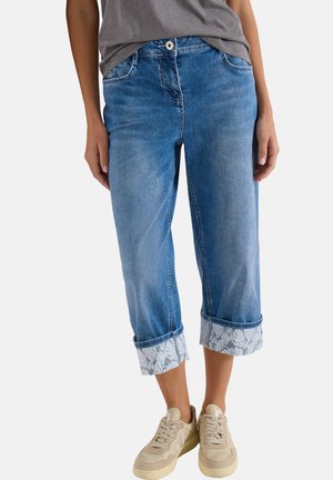 Femme portant un jean court bleu clair aux ourlets retournés à imprimé floral et des baskets beiges, debout devant un fond uni.