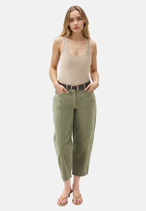 Junge Frau mit blonden Haaren, die ein beiges Tanktop, olivgrüne, hochgeschnittene Hosen mit einem schwarzen Gürtel und beige Absatzsandalen trägt und steht.