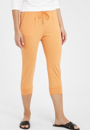 Pantalons capri orange en tissu doux, avec une taille élastique dotée d'un cordon de serrage et des ourlets retroussés. Portés avec des sandales noires à glissière.