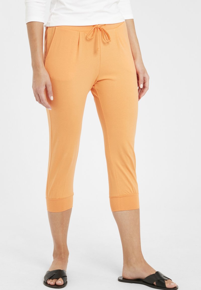Pantalons capri orange en tissu doux, avec une taille élastique dotée d'un cordon de serrage et des ourlets retroussés. Portés avec des sandales noires à glissière.