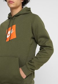 Olivgrön huvtröja med känguruficka, dragband och en orange rektangulär Nike-logotyp på bröstet. Mjuk bomullsblandning.