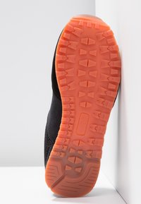 Chaussure noire avec semelle en caoutchouc orange montrant un motif de semelle détaillé, positionnée verticalement contre un fond blanc en angle.