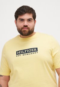 Gul bomull t-skjorte med en marineblå trykt logo "HILFIGER EST. MCMLXXXV" sentrert på brystet. Korte ermer, rund hals.