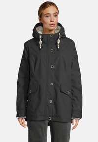 Vrouw in een zwarte hoodie met knopen aan de voorkant, klepzakken en witte trekkoorden, gecombineerd met donkergrijze broek.