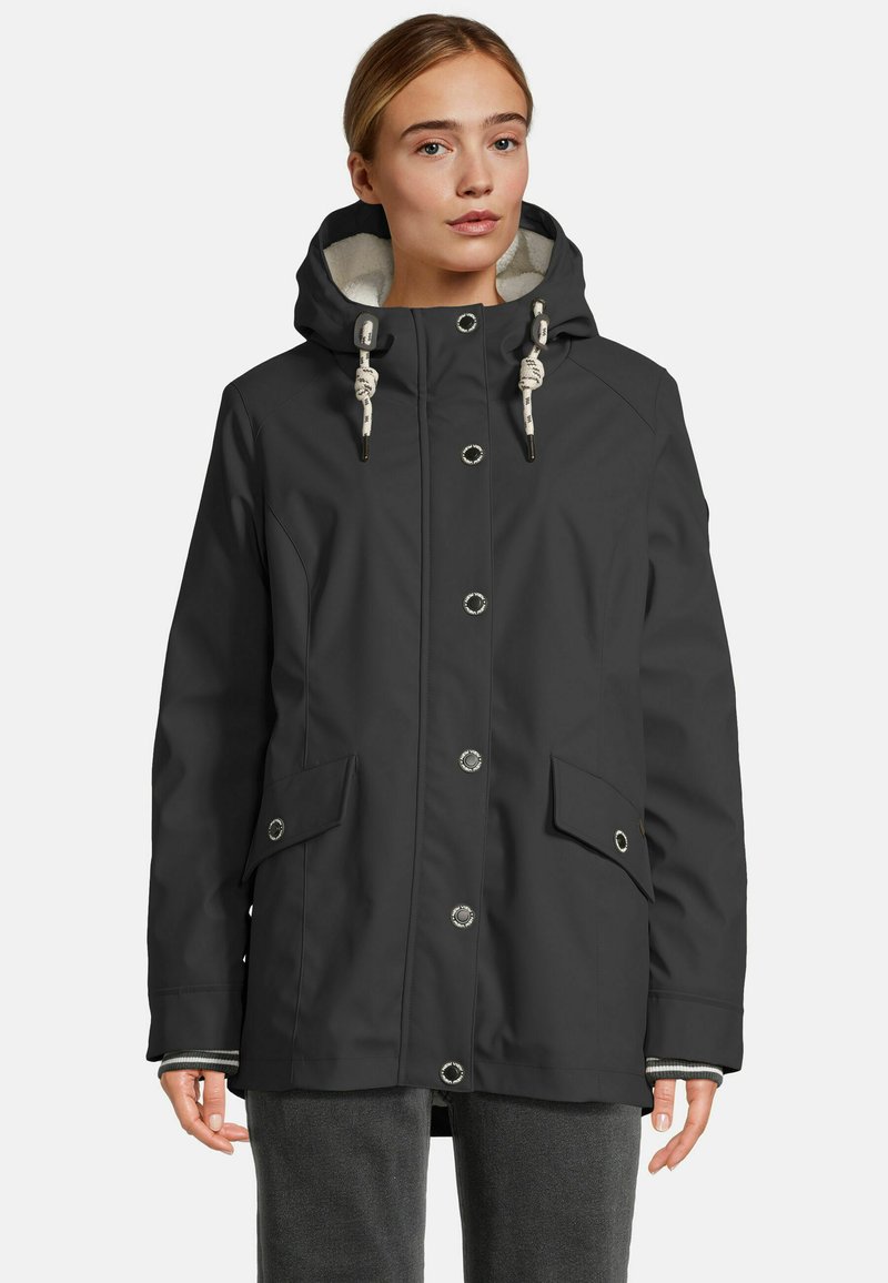 Vrouw in een zwarte hoodie met knopen aan de voorkant, klepzakken en witte trekkoorden, gecombineerd met donkergrijze broek.