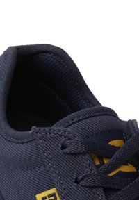 Navy canvas sneaker with yellow accents, featuring a rounded toe, textured upper, and traditional lacing system. Close-up view of shoe opening.