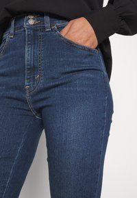 Mörkblå denimjeans med hög midja, försedda med två knappar, en framficka och synliga sömnadsdetaljer. Slät textur, åtsittande form.
