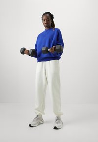 Felpa blu, pantaloni sportivi bianchi, che tiene manubri esagonali neri. Scarpe da ginnastica grigie con dettagli. Sfondo neutro.