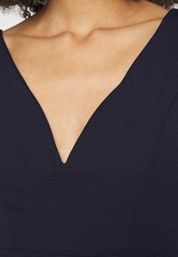 Abito navy con scollatura a V profonda, tessuto liscio e silhouette strutturata. I bordi della scollatura sono rifiniti in modo pulito.