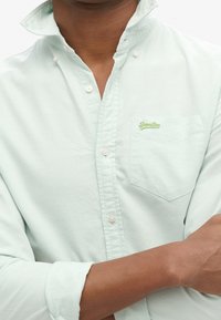 Lichtgroen katoenen overhemd met knoopsluiting, een kraag, één borstzak en geborduurd "SuperDry"-logo. De textuur oogt glad en lichtgewicht.