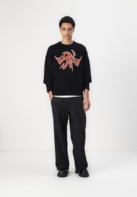 Sort sweater med ildmønsteret rød grafik, parret med løstsiddende grå ternede bukser og sorte chunky sko på en ensfarvet baggrund.