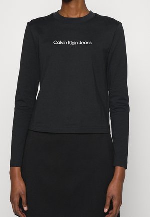 Person trägt ein schwarzes, langärmliges Croptop mit weißem Schriftzug „Calvin Klein Jeans“ in der Mitte der Brust.
