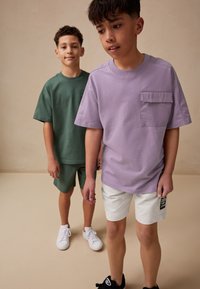 Dwóch chłopców ma na sobie codzienne stroje: jeden w zielonej, luźnej koszulce i szortach, drugi w lawendowej koszulce i białych szortach.