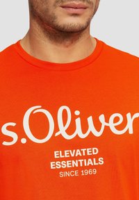 Oranžové bavlněné tričko s bílým nápisem "s.Oliver" a pod ním "ELEVATED ESSENTIALS" a "SINCE 1969", kulatý výstřih, hladká textura.